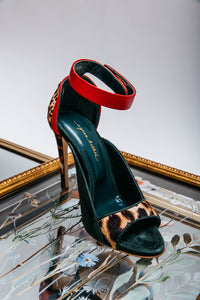 High-heeled sandal with red ankle strap and leopard print sole on a reflective surface.