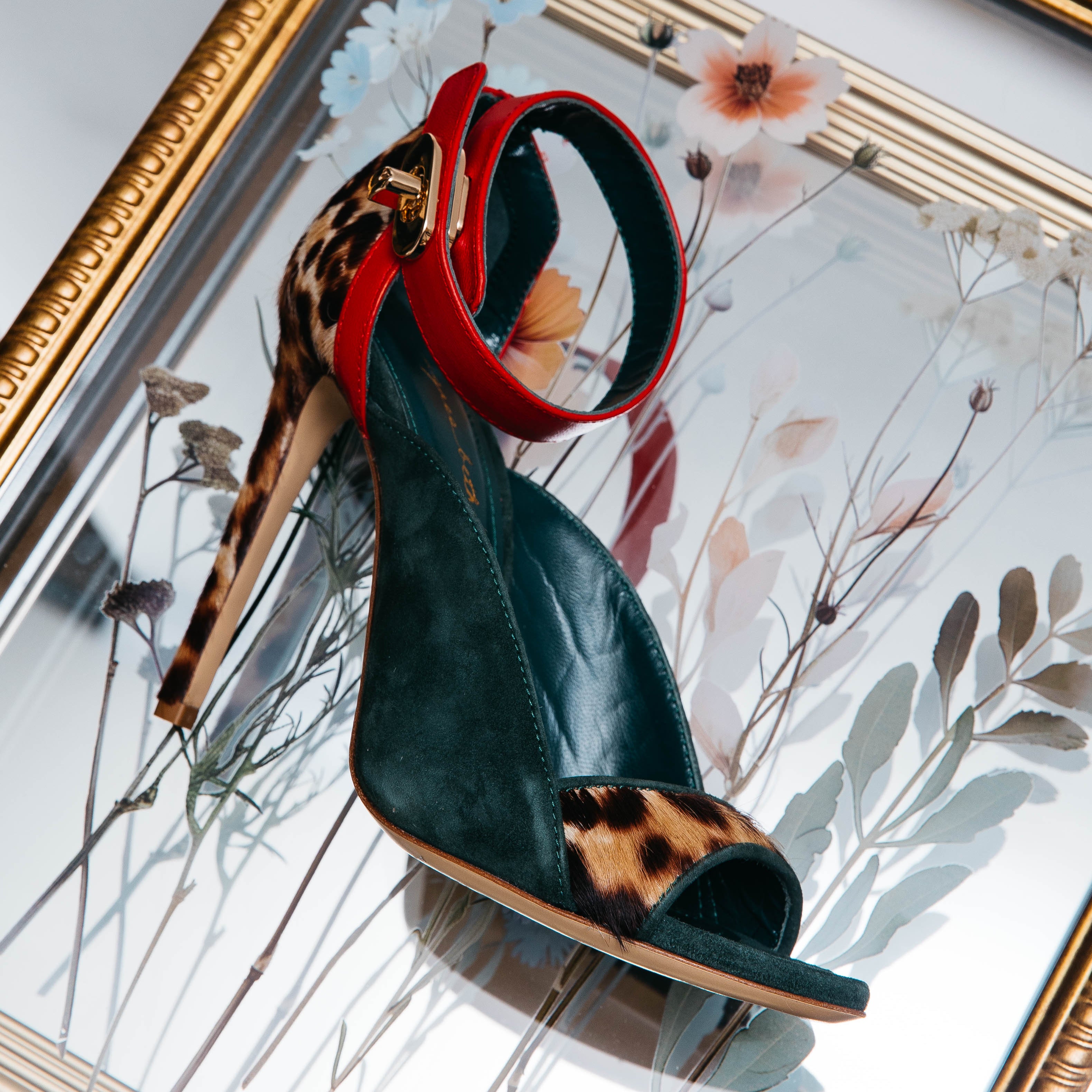 Green high-heeled shoe with red interior and leopard print sole in front of a floral painting.