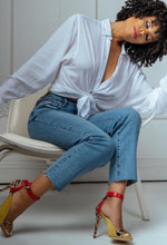 Load image into Gallery viewer, A woman with curly black hair sits on a white chair, wearing a white long-sleeve button-up shirt tied at the waist, blue cropped jeans, and yellow high-heeled sandals with red ankle straps and leopard print detailing.
