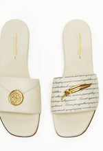 Load image into Gallery viewer, A pair of beige-colored sandals, one featuring a gold seal pendant and the other with a quill pin detail.
