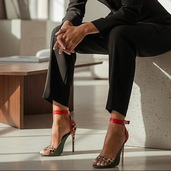 Person wearing a black suit and green, leopard print and red high heels sitting on a concrete bench.