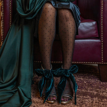 Person wearing a green velvet sandals with a bow polka dot tights sitting on a red leather chair.